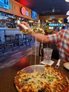 man's hand lifting piece of cheesy pizza out of pan in a restaurant with a bar behind him