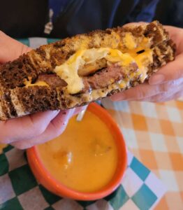 Close-up of person holding sandwich with bowl of soup in the background