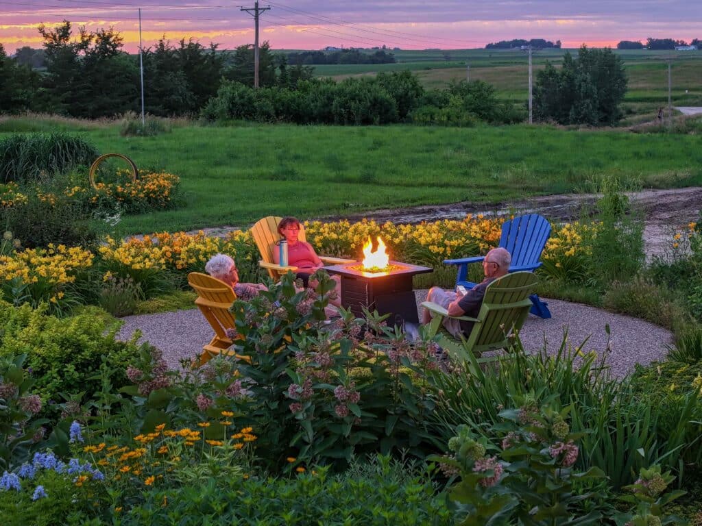 3 people sitting in chairs around a fire pit in a garden with a sunset behind them 